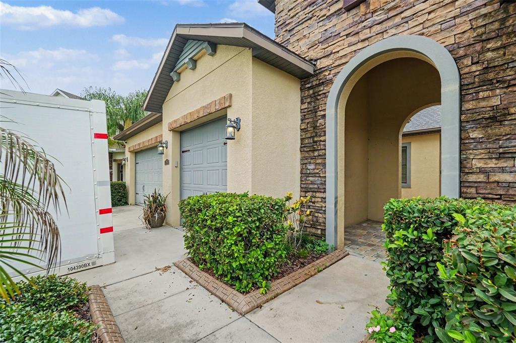 A stone-accented house exterior features a beige garage, a covered entryway, and landscaped bushes — 31957 MARDELLE PLACE, WESLEY CHAPEL, FL