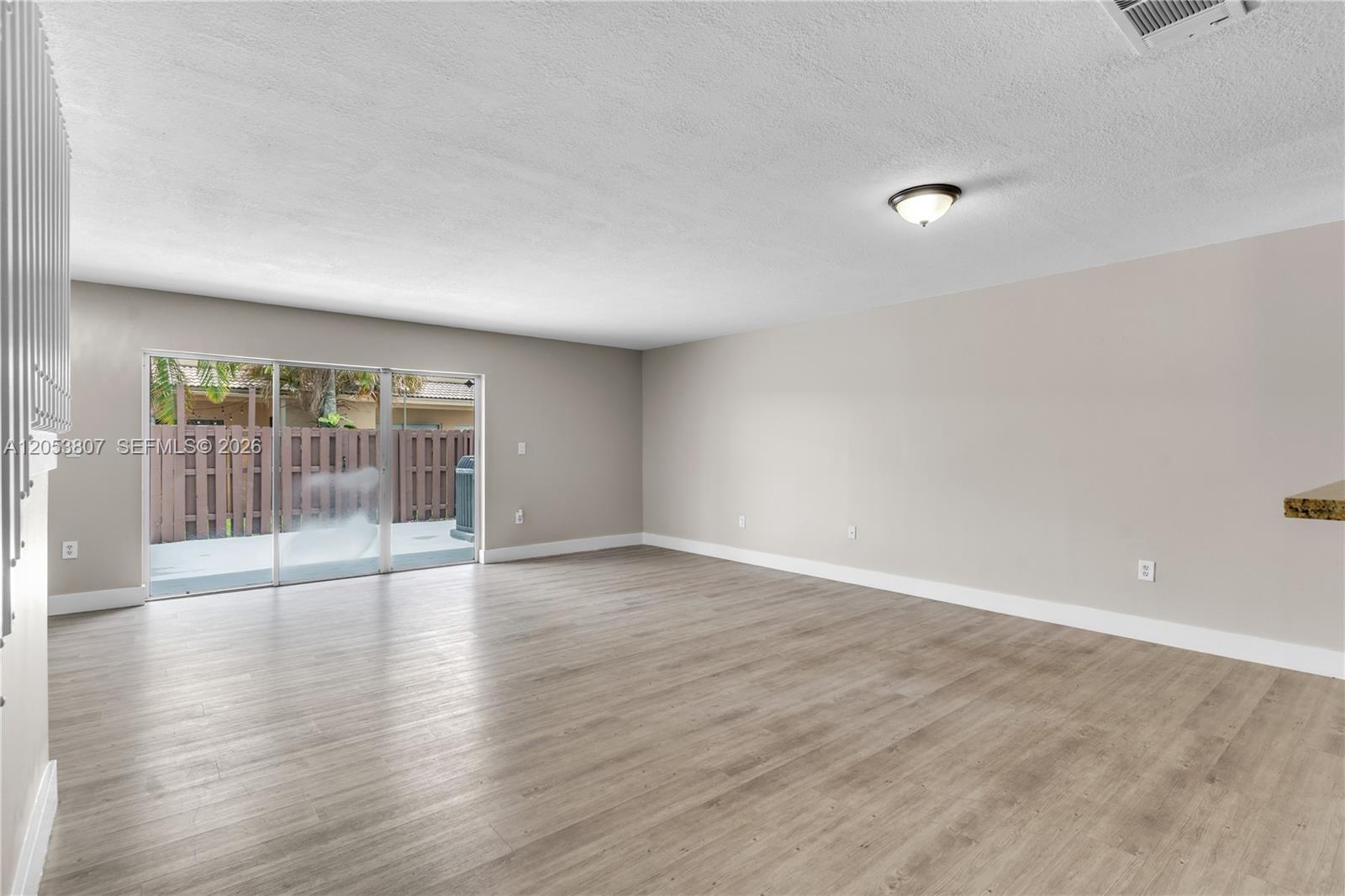 An empty living room features light wood-look flooring, beige walls, and sliding glass patio doors — 7463 SW 164th Ave, Miami FL 33193, MIAMI, FL