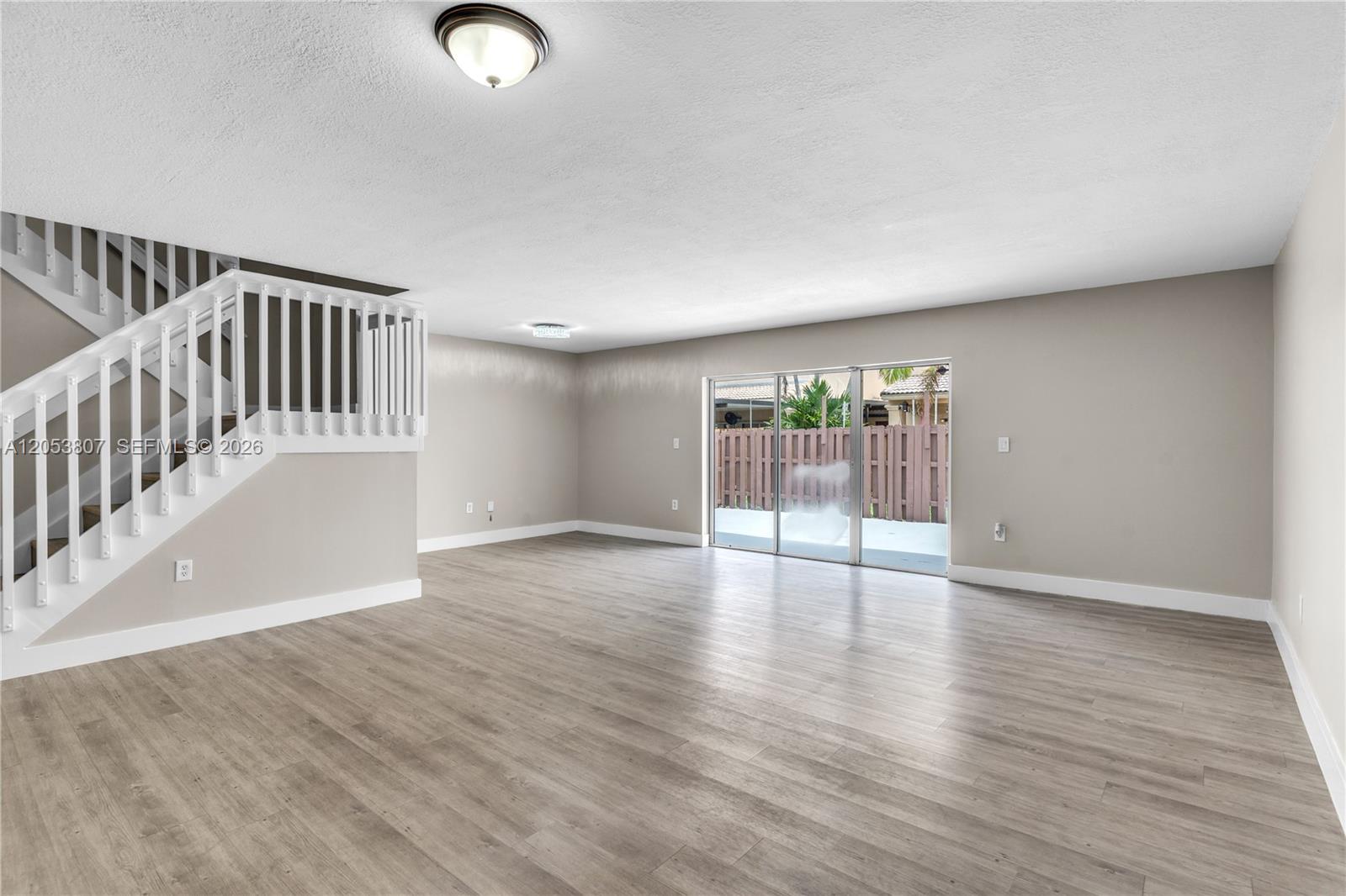 An empty living room features light wood-look flooring, a staircase with white railings, and sliding doors — 7463 SW 164th Ave, Miami FL 33193, MIAMI, FL