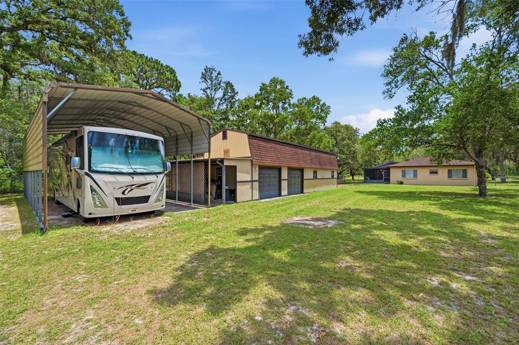 A large RV parked under a metal carport next to a detached garage and house — 15041 CAMROSE AVENUE, SPRING HILL, FL