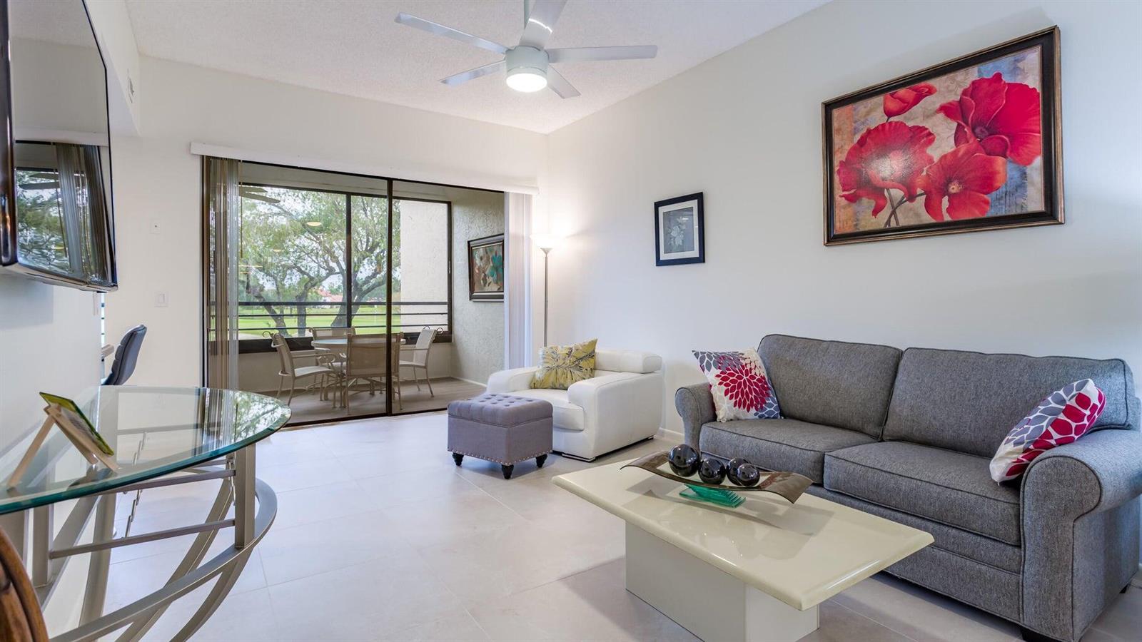 A living room features a gray sofa, white armchair, glass coffee table, and a balcony view — 7460 La Paz Boulevard # 208, Boca Raton FL 33433, BOCA RATON, FL