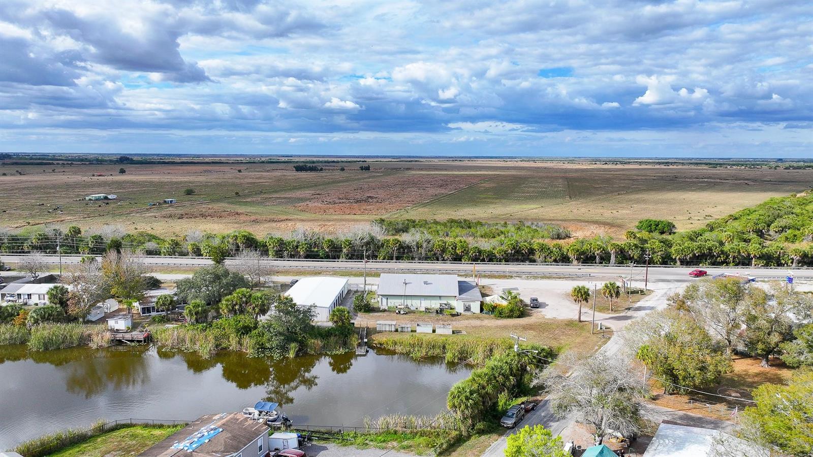 An aerial view shows a pond, a commercial building, and a highway bordering open fields — 1110 Buckhead Ridge Road, Okeechobee FL 34974, OKEECHOBEE, FL