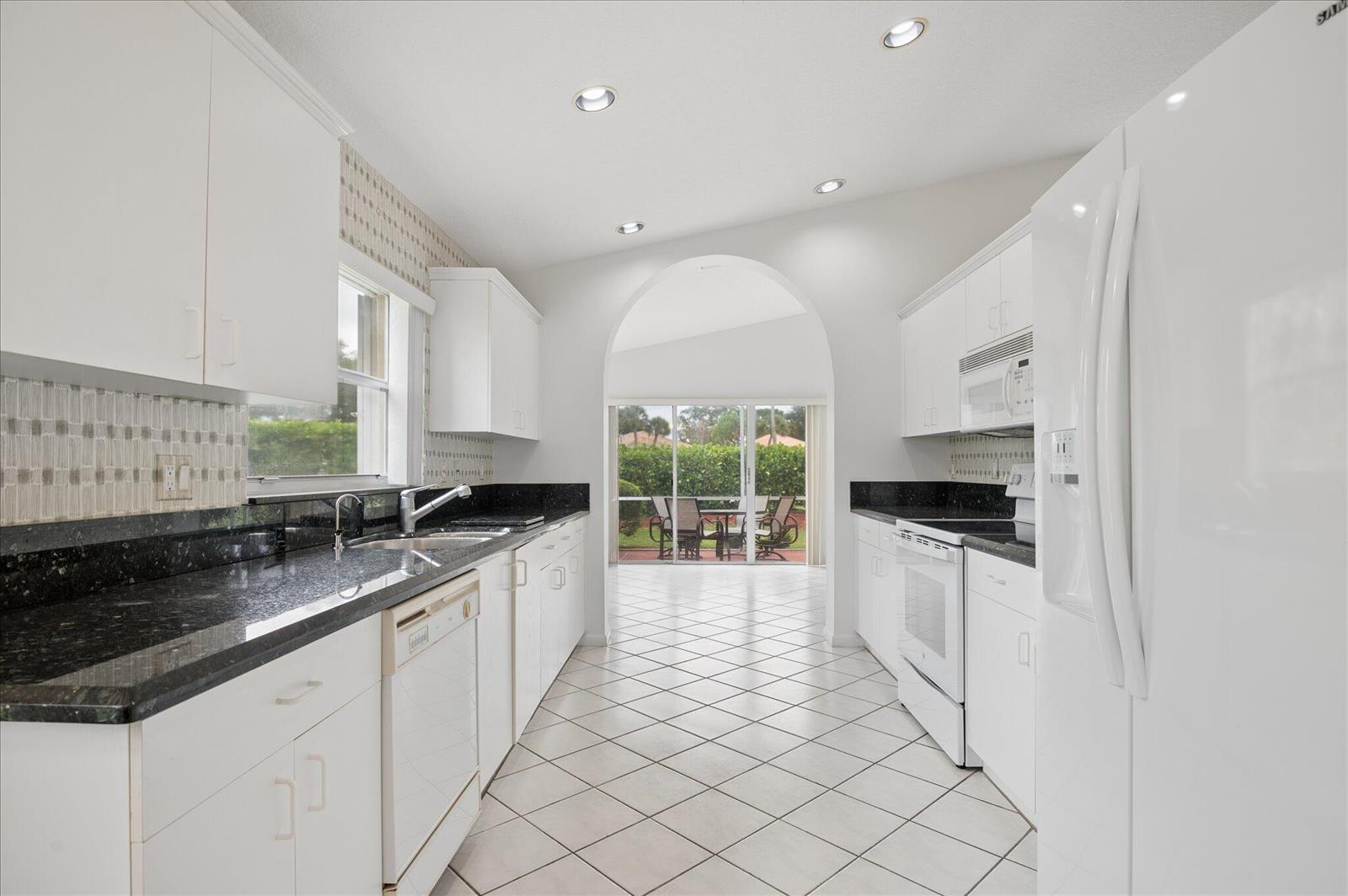 A galley kitchen with white cabinets, black countertops, and a tiled floor leads to a patio — 10884 Royal Caribbean Circle, Boynton Beach FL 33437, BOYNTON BEACH, FL