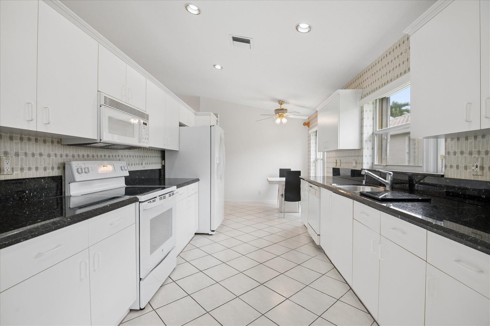 A galley kitchen features white cabinets, black countertops, white appliances, and light-colored tile flooring — 10884 Royal Caribbean Circle, Boynton Beach FL 33437, BOYNTON BEACH, FL