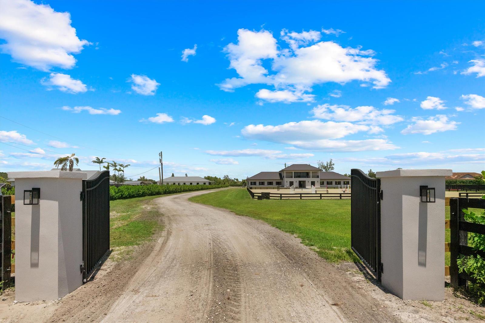 A dirt driveway leads through an open gate toward a large house and equestrian arena — 16175 Deer Path Lane, Wellington FL 33470, WELLINGTON, FL