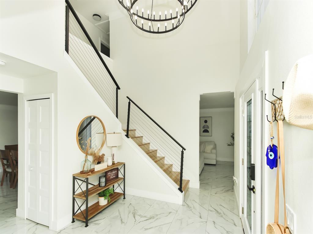 A bright foyer features a staircase with cable railings, marble-look tile flooring, and a chandelier — 7561 LAKE ANDREA CIRCLE, MOUNT DORA, FL