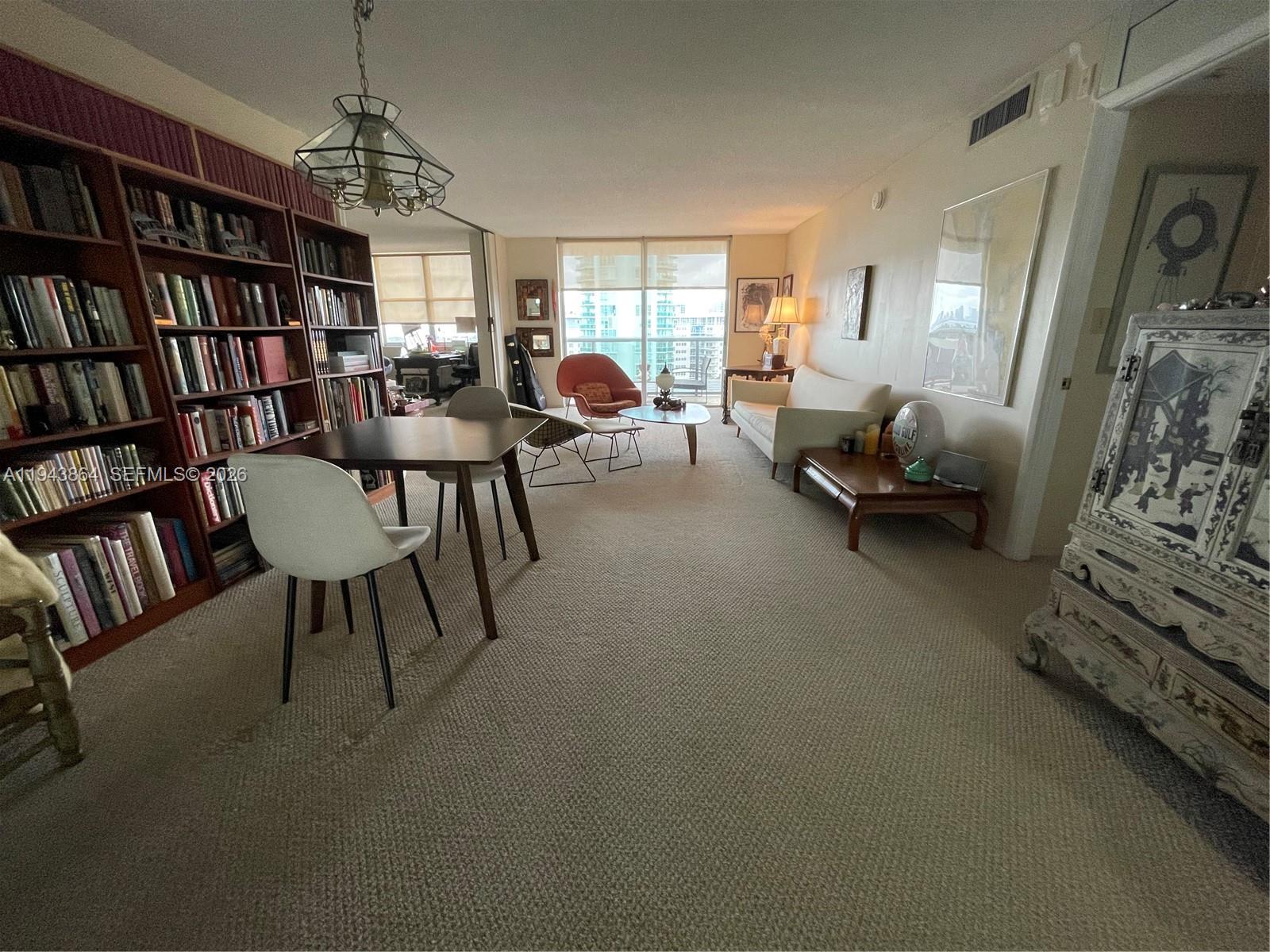 A living and dining area features a large bookshelf, carpeted floors, and floor-to-ceiling windows — 6039 Collins Ave # 1232, Miami Beach FL 33140, MIAMI BEACH, FL