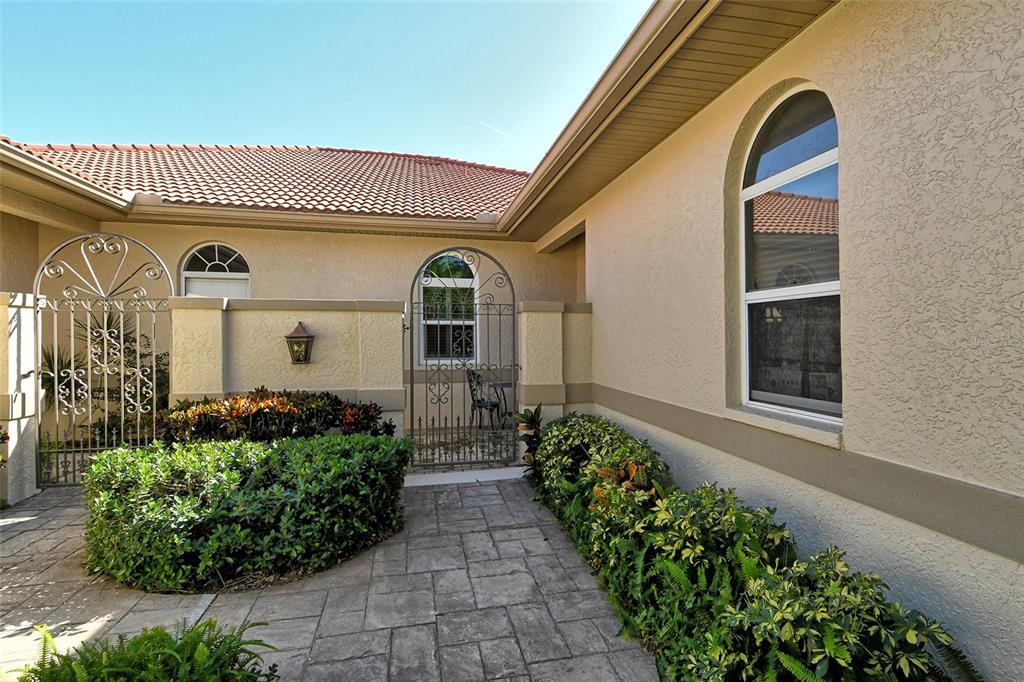 A paved walkway leads to a gated courtyard entrance with stucco walls and arched windows — 3416 W CHELMSFORD COURT UNIT 2, SARASOTA, FL