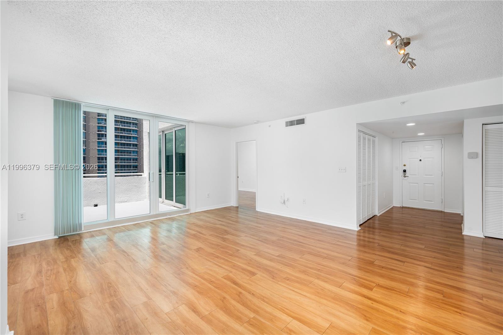 An empty living room features light wood-look flooring, white walls, and a sliding glass door — 1111 Brickell Bay Dr # 211, Miami FL 33131, MIAMI, FL