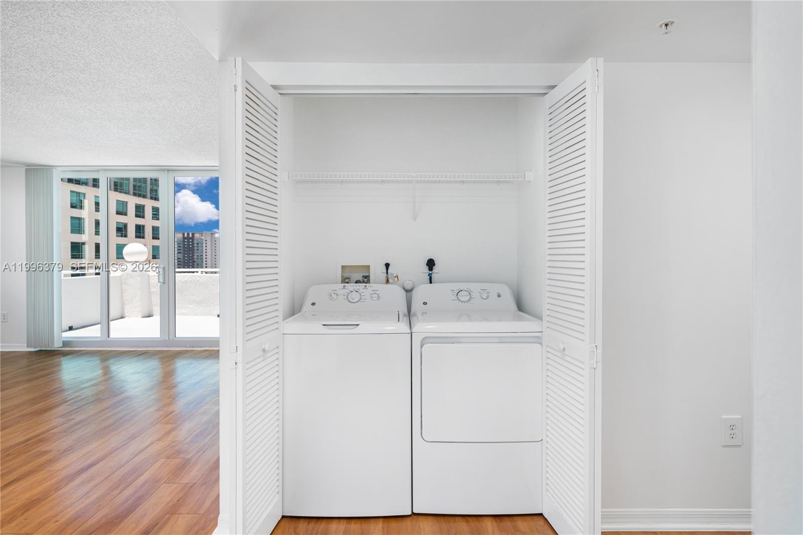A white laundry closet with a washer and dryer sits beside a room with hardwood floors — 1111 Brickell Bay Dr # 211, Miami FL 33131, MIAMI, FL