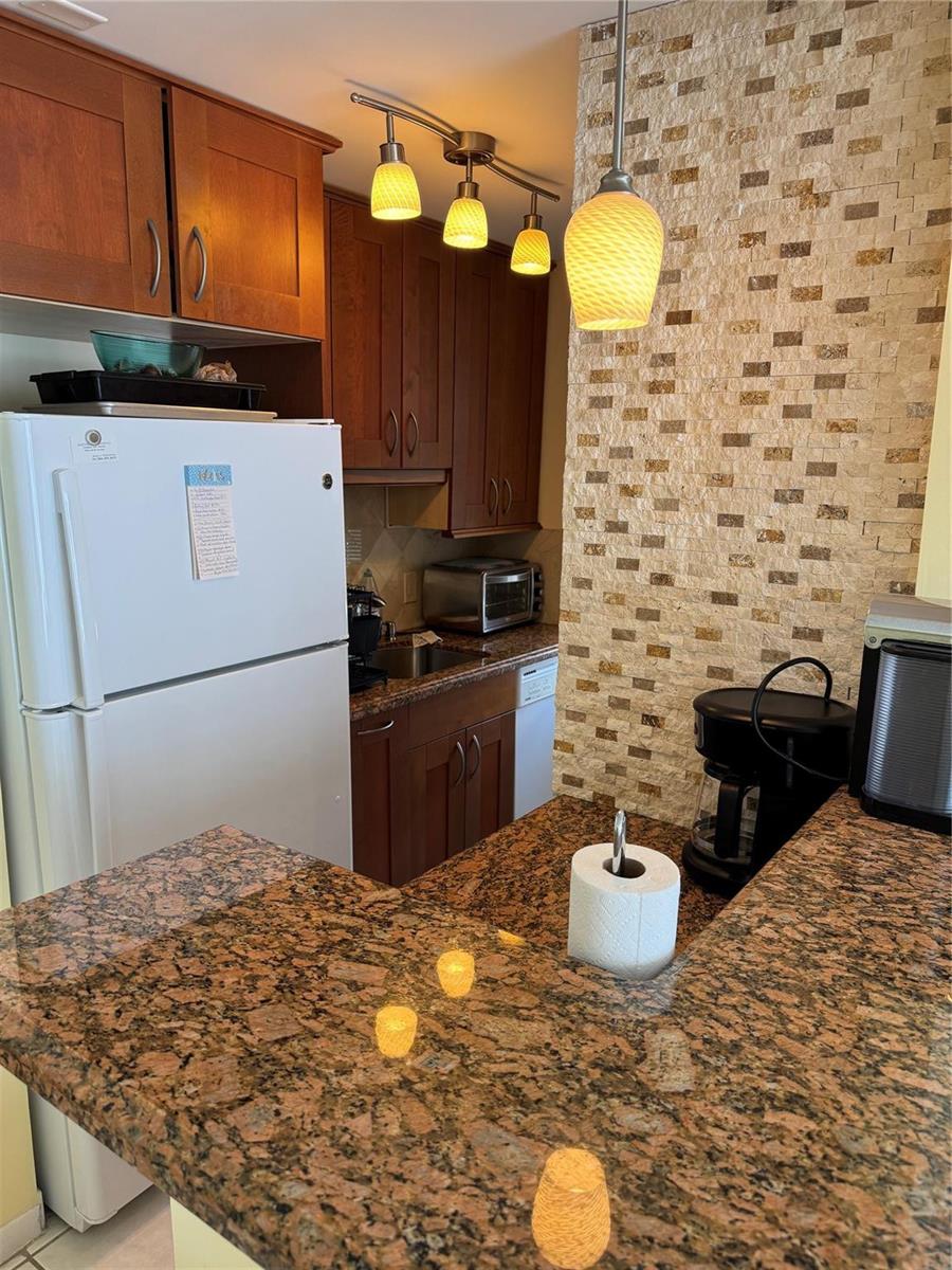 A kitchen features wood cabinets, granite countertops, a stone-tiled accent wall, and pendant lighting — 111 Briny Avenue # 11, Pompano Beach FL 33062, POMPANO BEACH, FL