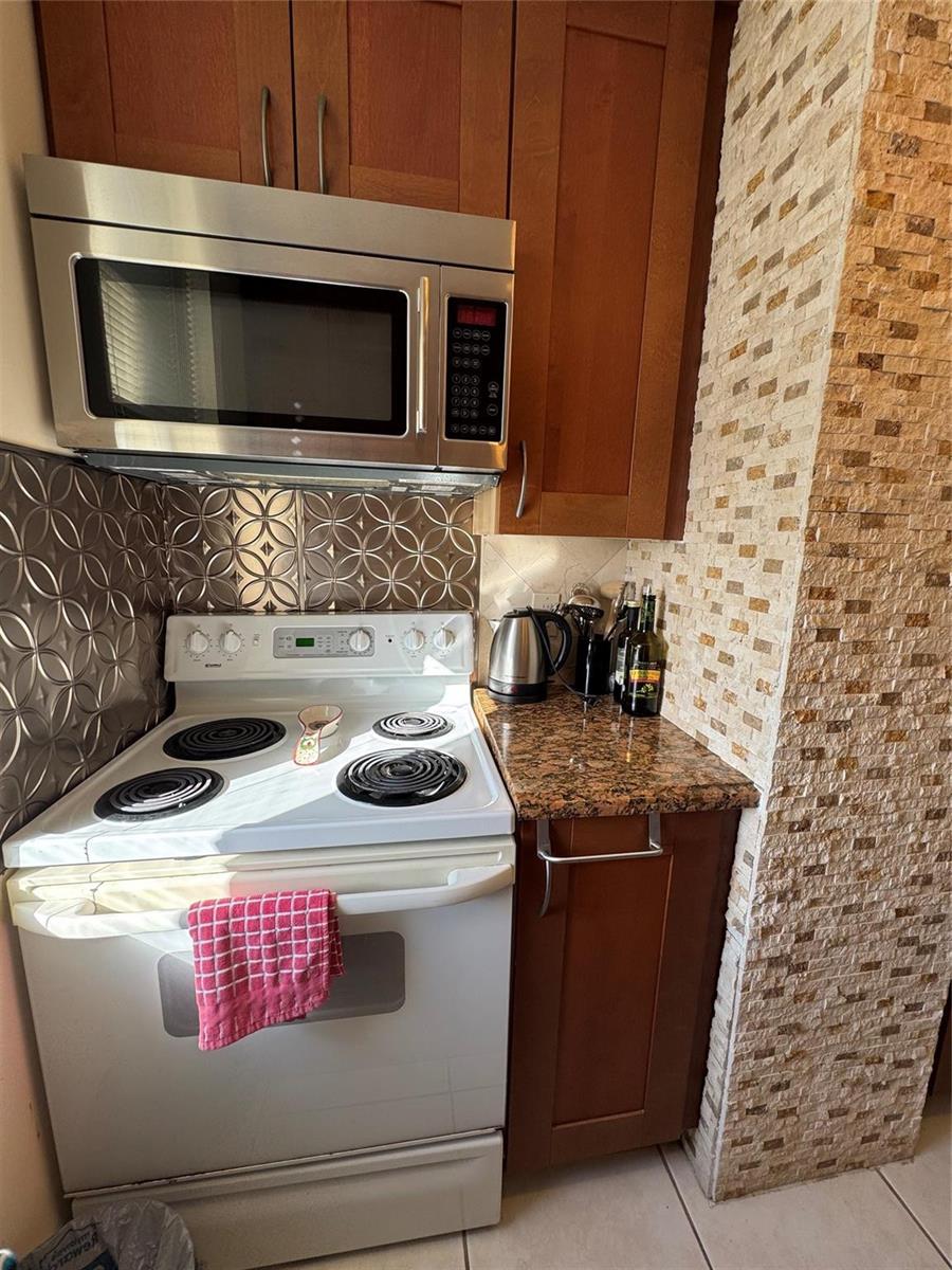 A kitchen features a white electric stove, stainless steel microwave, wood cabinets, and stone backsplash — 111 Briny Avenue # 11, Pompano Beach FL 33062, POMPANO BEACH, FL