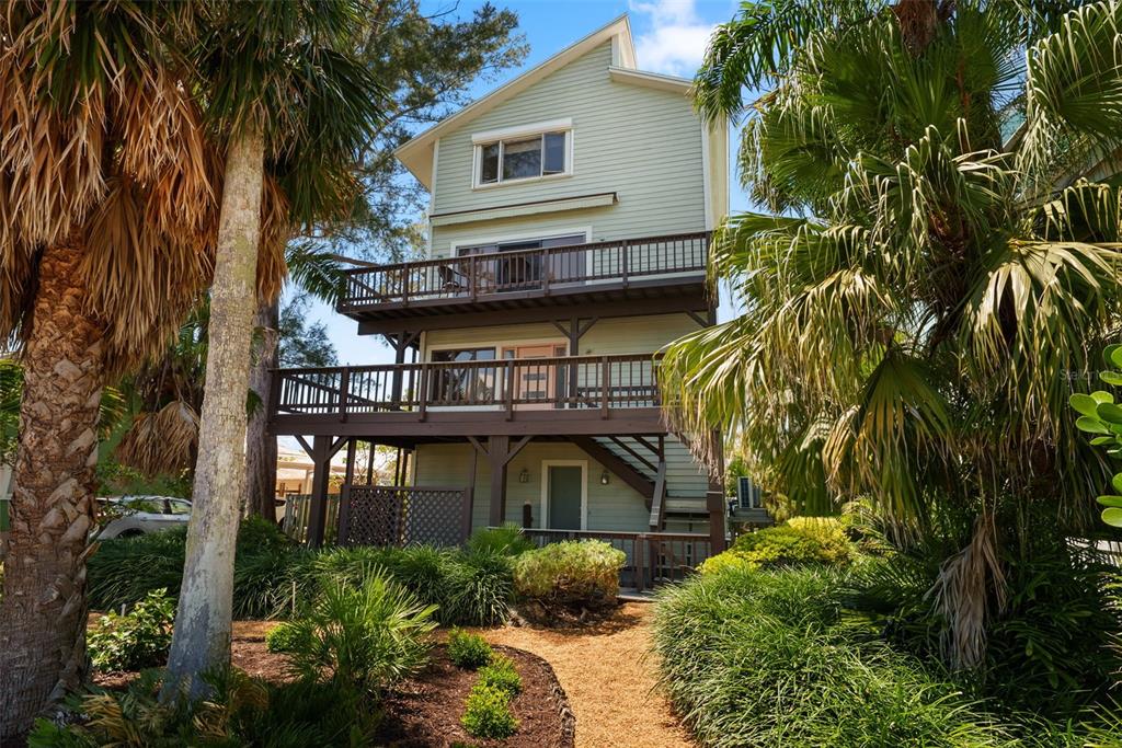 A three-story light green house with two wooden decks surrounded by lush tropical landscaping — 1304 PASS A GRILLE WAY, ST PETE BEACH, FL