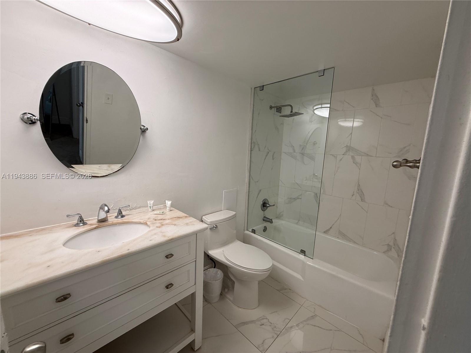A bathroom features a white vanity, a round mirror, a toilet, and a glass-enclosed tub — 5101 Collins Ave # 12T, Miami Beach FL 33140, MIAMI BEACH, FL