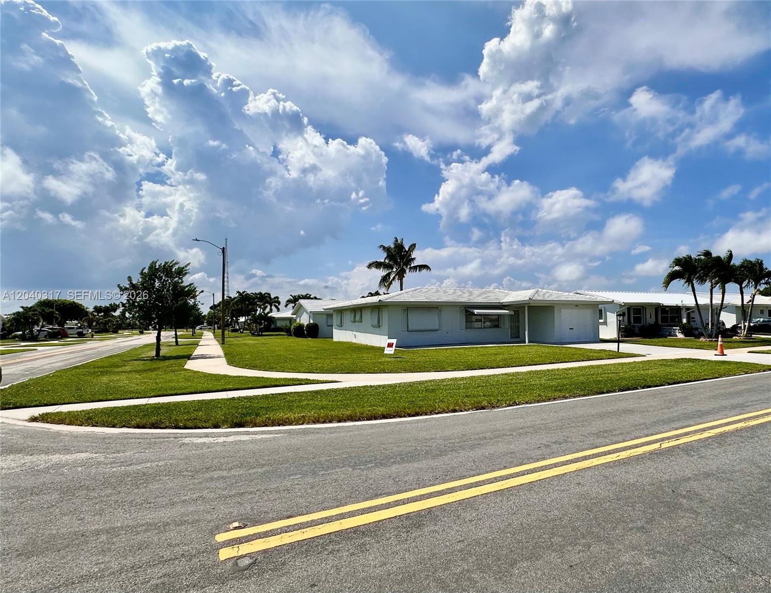 A light-colored single-story house with a lawn sits on a corner lot under clouds — BOYNTON BEACH, FL