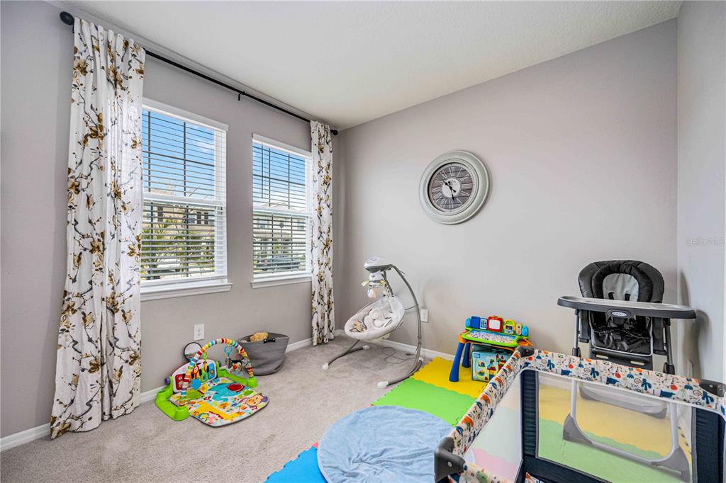 A nursery with gray walls, carpeted flooring, two windows with patterned curtains, and baby equipment — 5329 HALEWOOD COURT, BRADENTON, FL