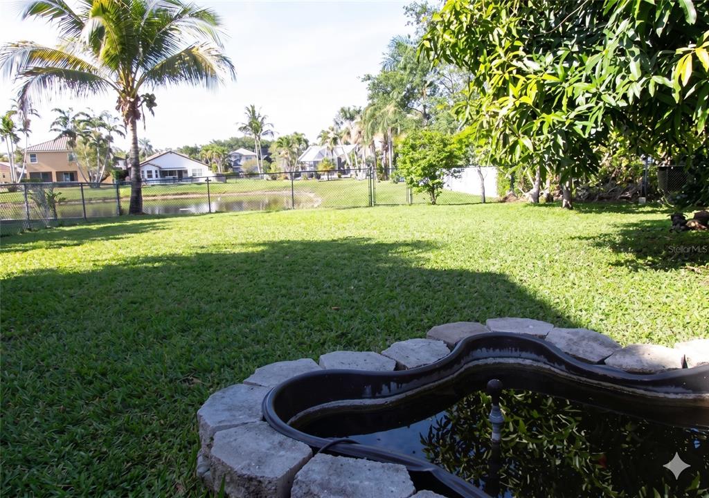 A backyard features a stone-bordered pond in the foreground, a grassy lawn, and palm trees — 11483 BEACON POINTE LANE, WELLINGTON, FL