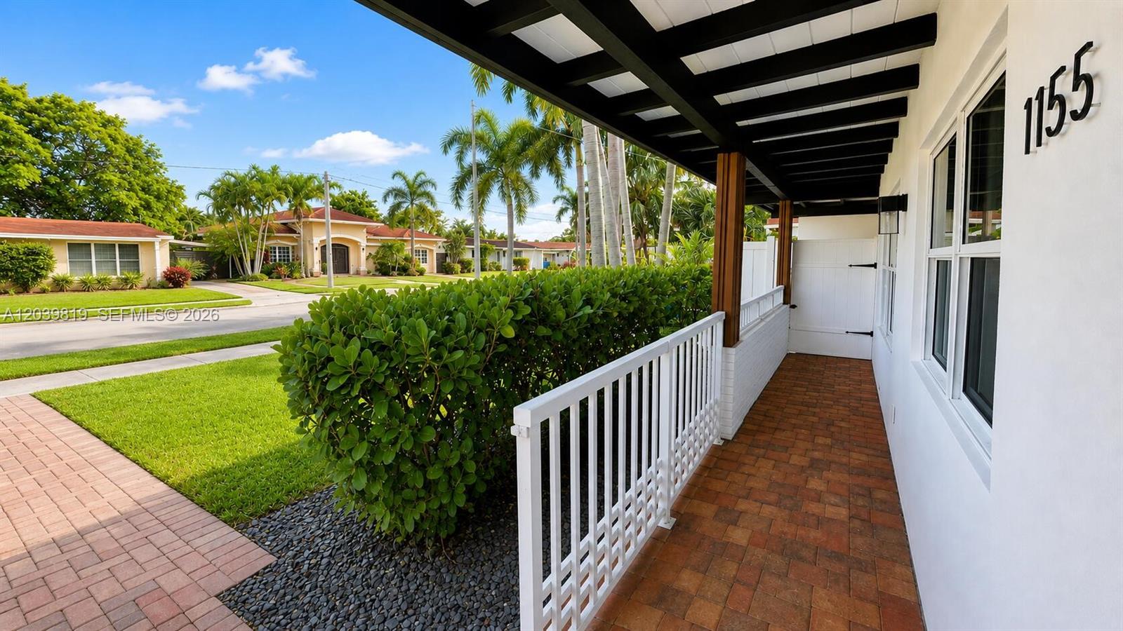 A covered brick porch with a white railing overlooks a lawn and palm trees — 1155 Hayes St, Hollywood FL 33019, HOLLYWOOD, FL