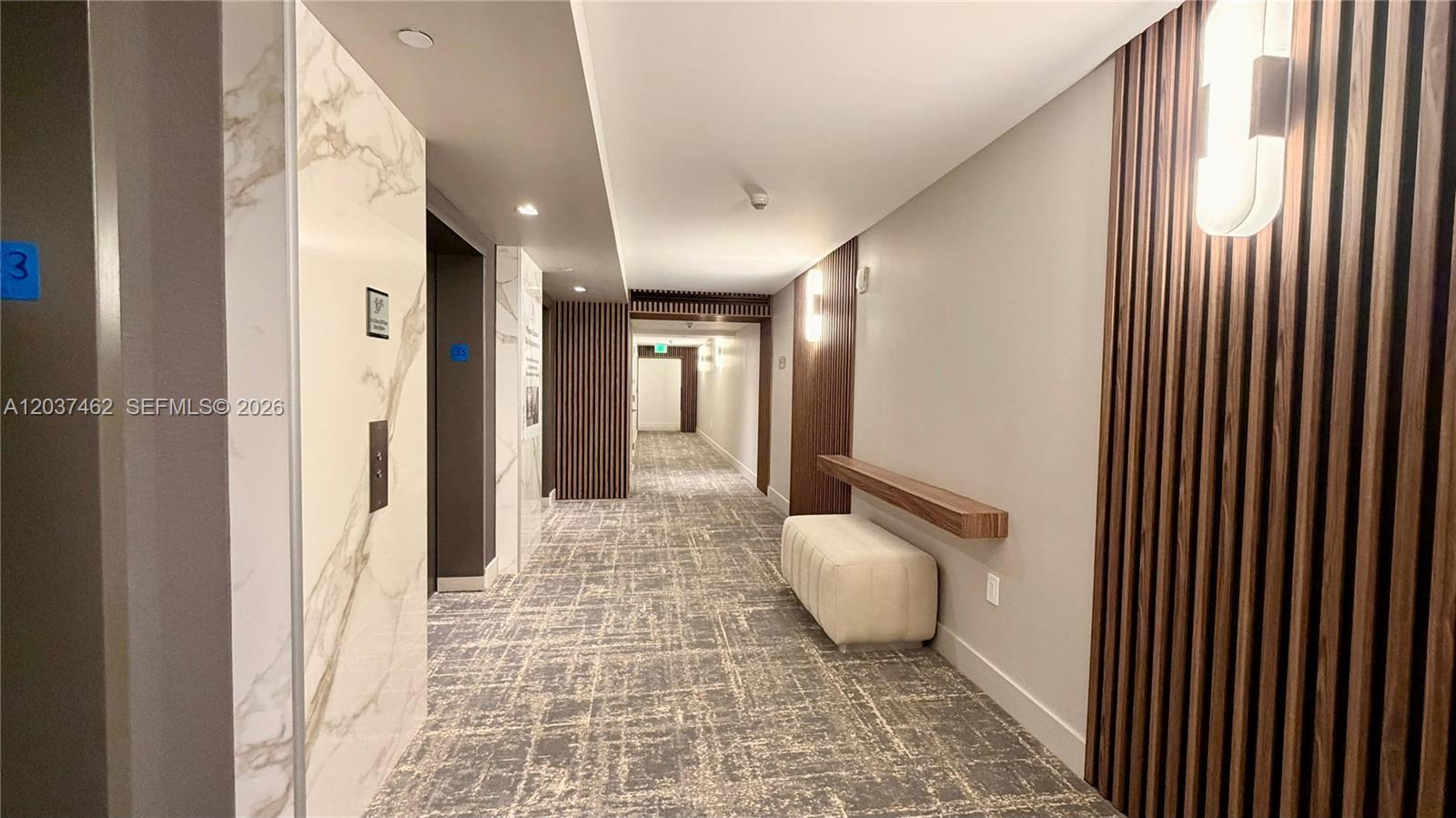A modern hallway features marble elevator surrounds, wood-slat wall paneling, and a cushioned bench — 6365 Collins Ave # 2201, Miami Beach FL 33141, MIAMI BEACH, FL