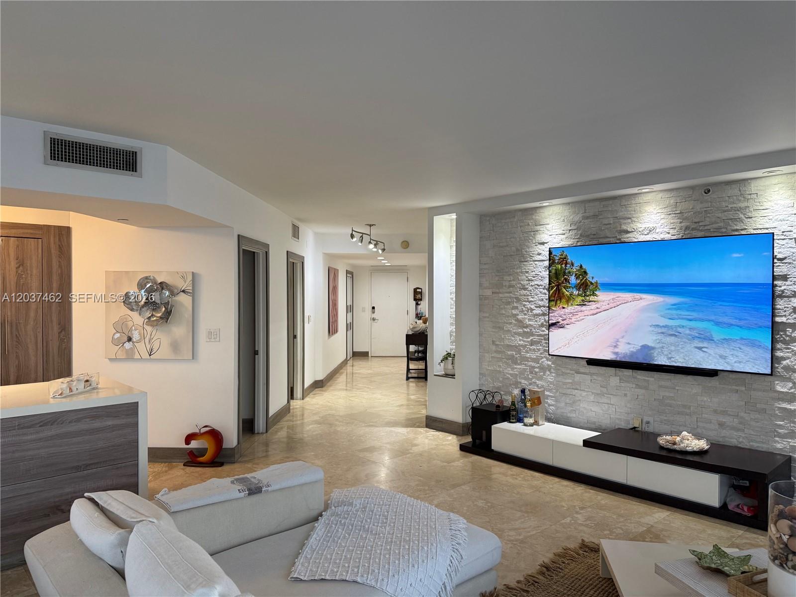 A living room features a stone accent wall with a large television and tile flooring — 6365 Collins Ave # 2201, Miami Beach FL 33141, MIAMI BEACH, FL