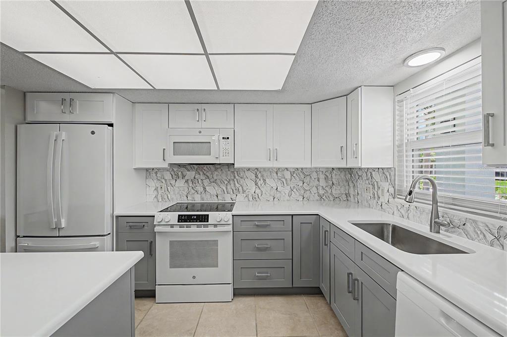 A kitchen features white upper cabinets, gray lower cabinets, white countertops, and a marble backsplash — 9 FORBES PLACE UNIT 108, DUNEDIN, FL
