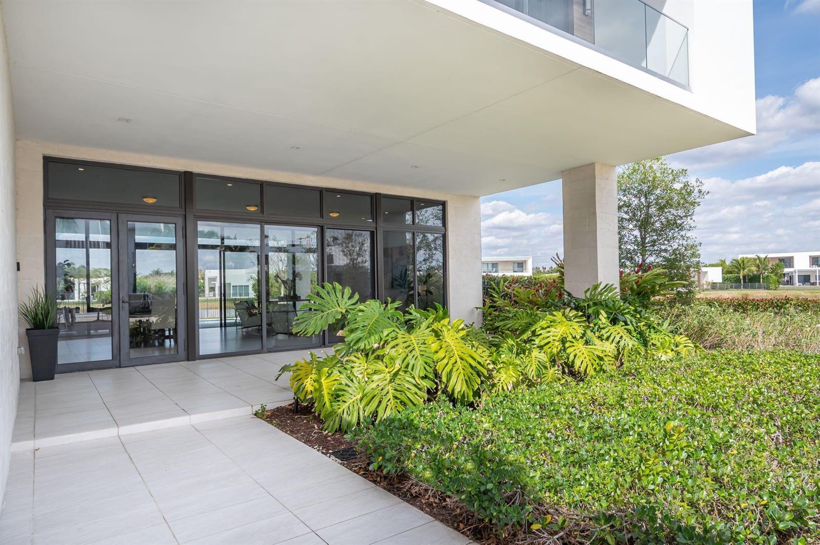 A modern home exterior features a tiled patio, large glass doors, and lush tropical landscaping — 16553 Botaniko Drive S, Weston FL 33326, WESTON, FL