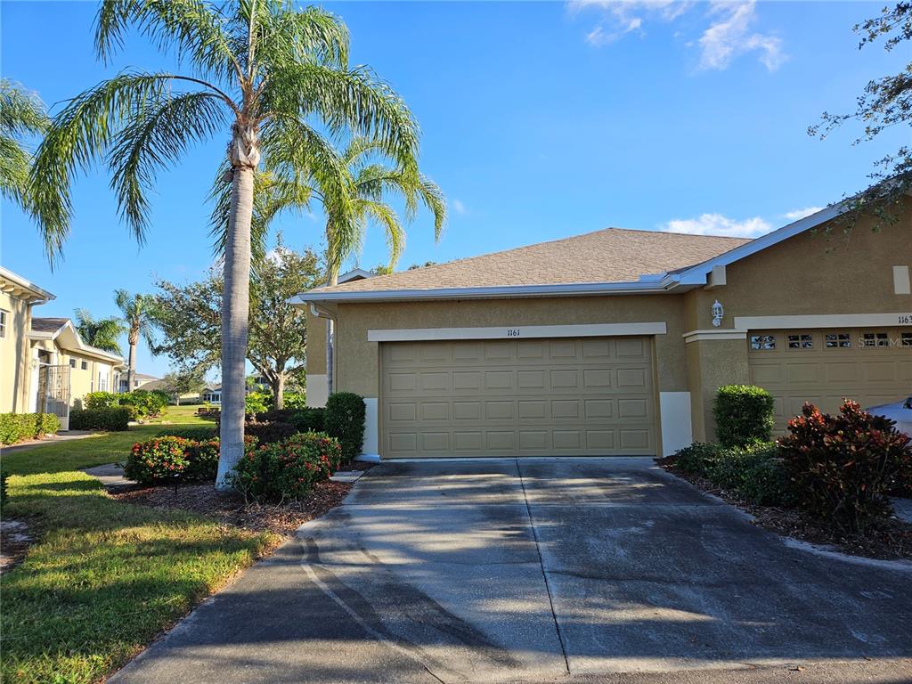 A tan house exterior features a two-car garage, a palm tree, and a concrete driveway — 1161 JAMESON GREENS DRIVE, SUN CITY CENTER, FL
