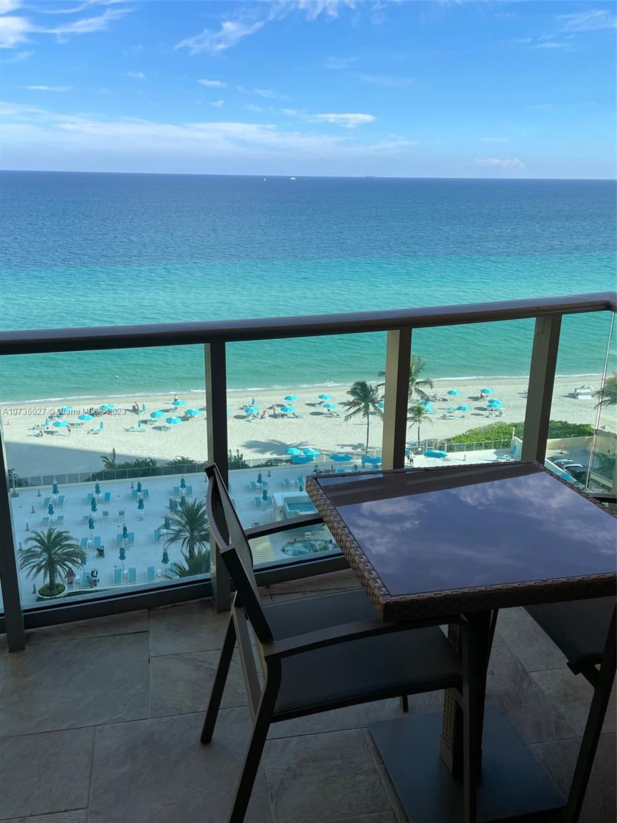 A balcony with a table and chairs overlooks a beach with blue umbrellas and ocean — 2501 S OCEAN DR, Hollywood FL 33019, HOLLYWOOD, FL