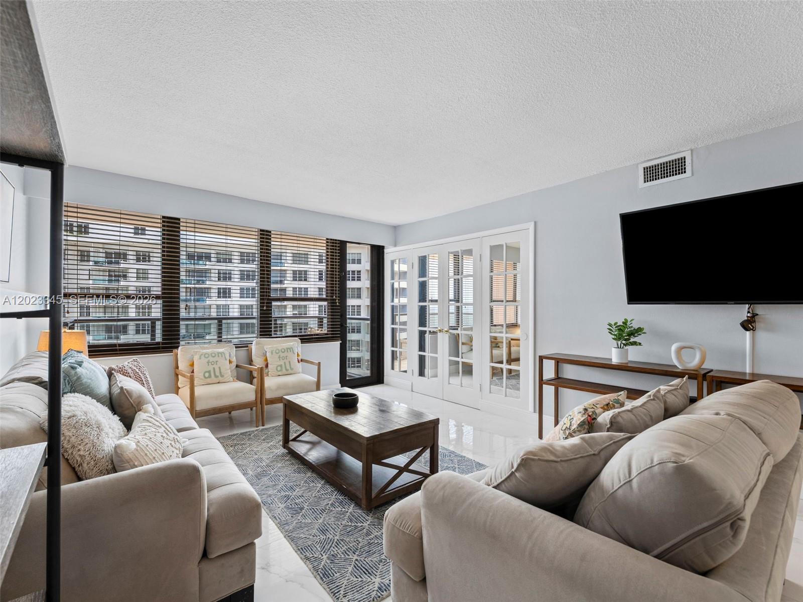 A living room features light-colored sofas, a wooden coffee table, and large windows with blinds — 5225 Collins Ave # 1509, Miami Beach FL 33140, MIAMI BEACH, FL