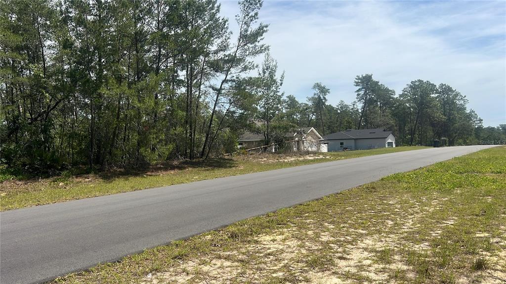 A paved road runs past a wooded lot with a house visible in the background — TBD MARION OAKS GOLF WAY, OCALA, FL