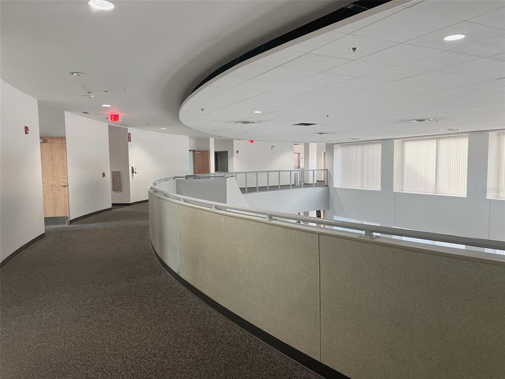 A curved carpeted hallway overlooks an open atrium with large windows and a metal railing — 800 CELEBRATION AVENUE UNIT 201-F, CELEBRATION, FL