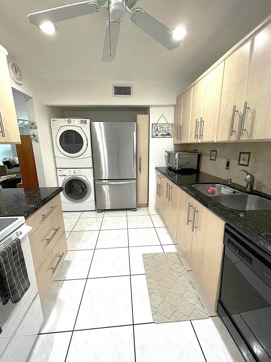 A kitchen with light wood cabinets, black countertops, white tile floors, and a stacked laundry unit — 2431 Del Aire Boulevard # d, Delray Beach FL 33445, DELRAY BEACH, FL