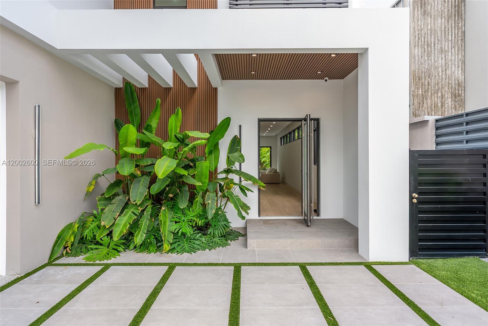 A modern home entrance features a paved walkway, lush tropical plants, and a wood-paneled ceiling — 260 NW 47th St, Miami FL 33127, MIAMI, FL
