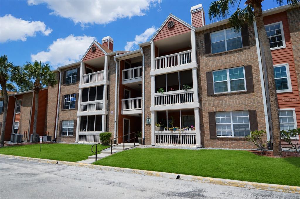 A three-story brick apartment building features screened balconies and a grassy lawn under a blue sky — 10200 GANDY BOULEVARD N UNIT 1313, ST PETERSBURG, FL