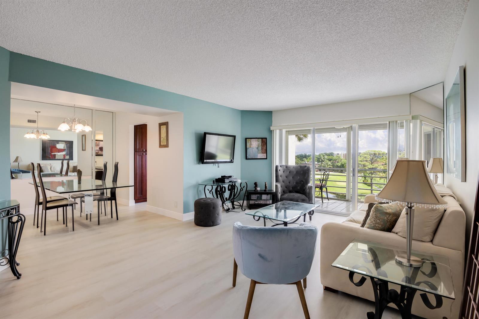 A living and dining area with light wood floors, blue accent walls, and a balcony view — 3593 Birdie Drive # 507, Lake Worth FL 33467, LAKE WORTH, FL