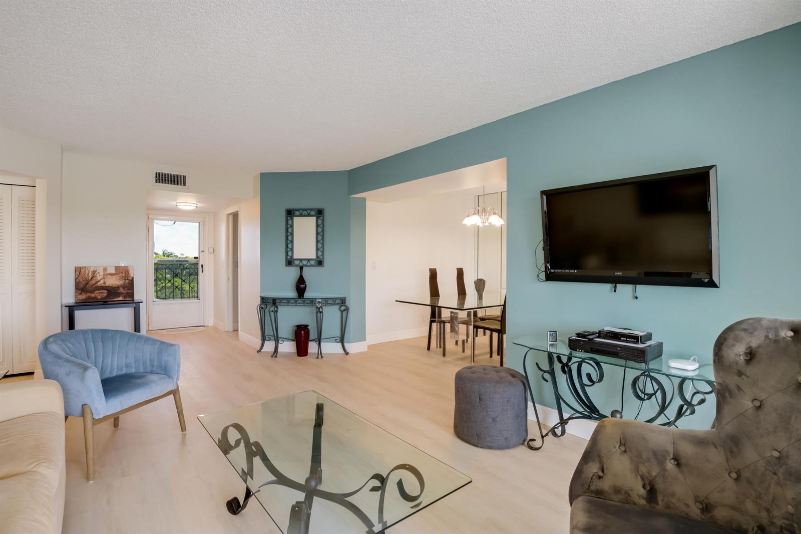 A living room with light wood floors, blue accent walls, a television, and glass tables — 3593 Birdie Drive # 507, Lake Worth FL 33467, LAKE WORTH, FL