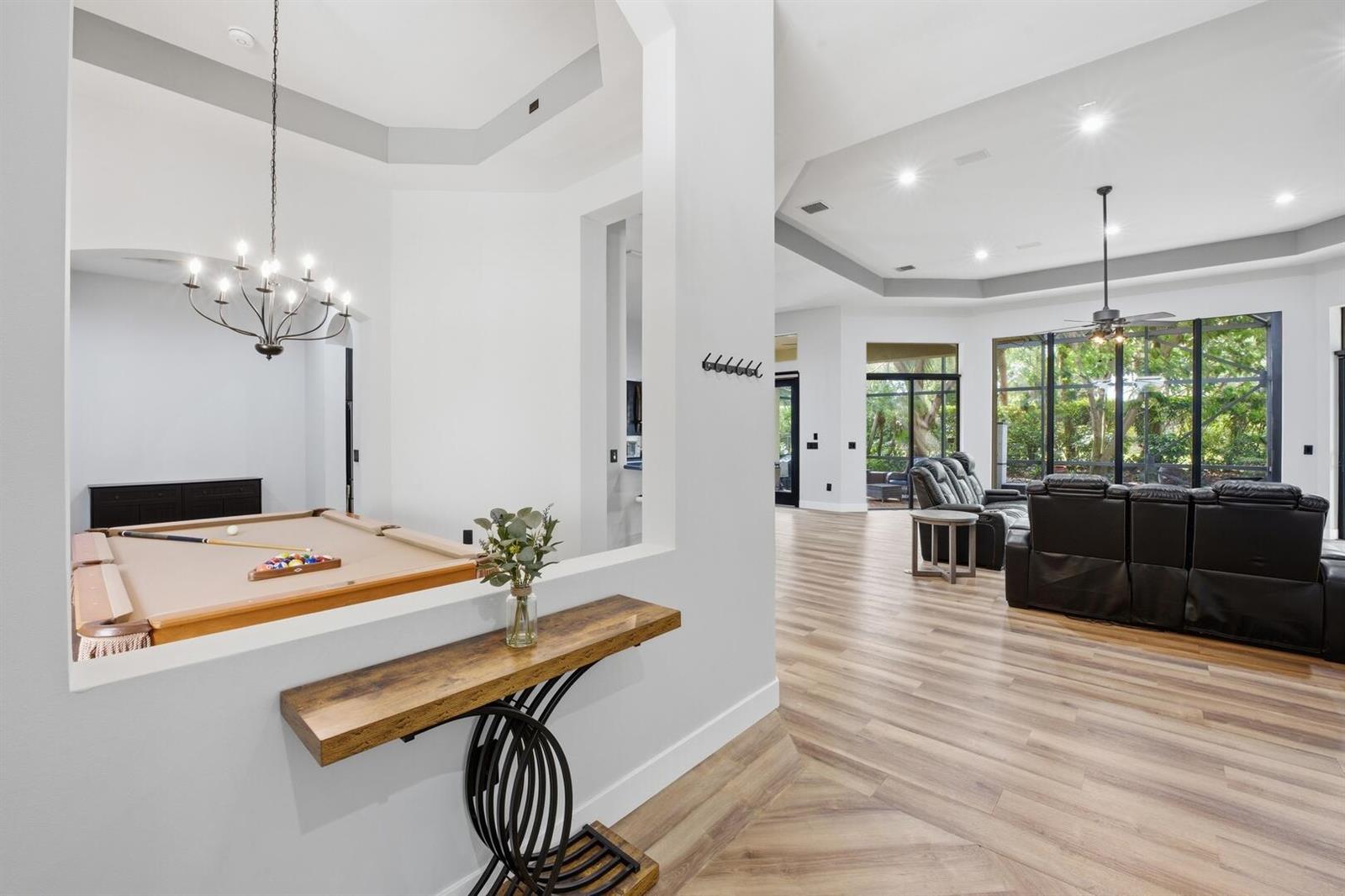 A living room with a pool table, wood-look flooring, and large windows overlooking trees — 6983 Antinori Lane, Boynton Beach FL 33437, BOYNTON BEACH, FL