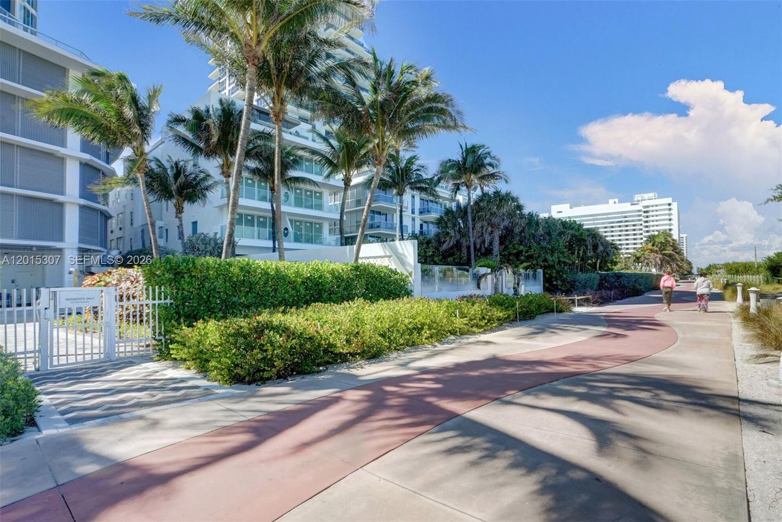 A paved walkway leads past a white fence, lush landscaping, and a modern residential building — 6515 Collins Ave # 1107, Miami Beach FL 33141, MIAMI BEACH, FL