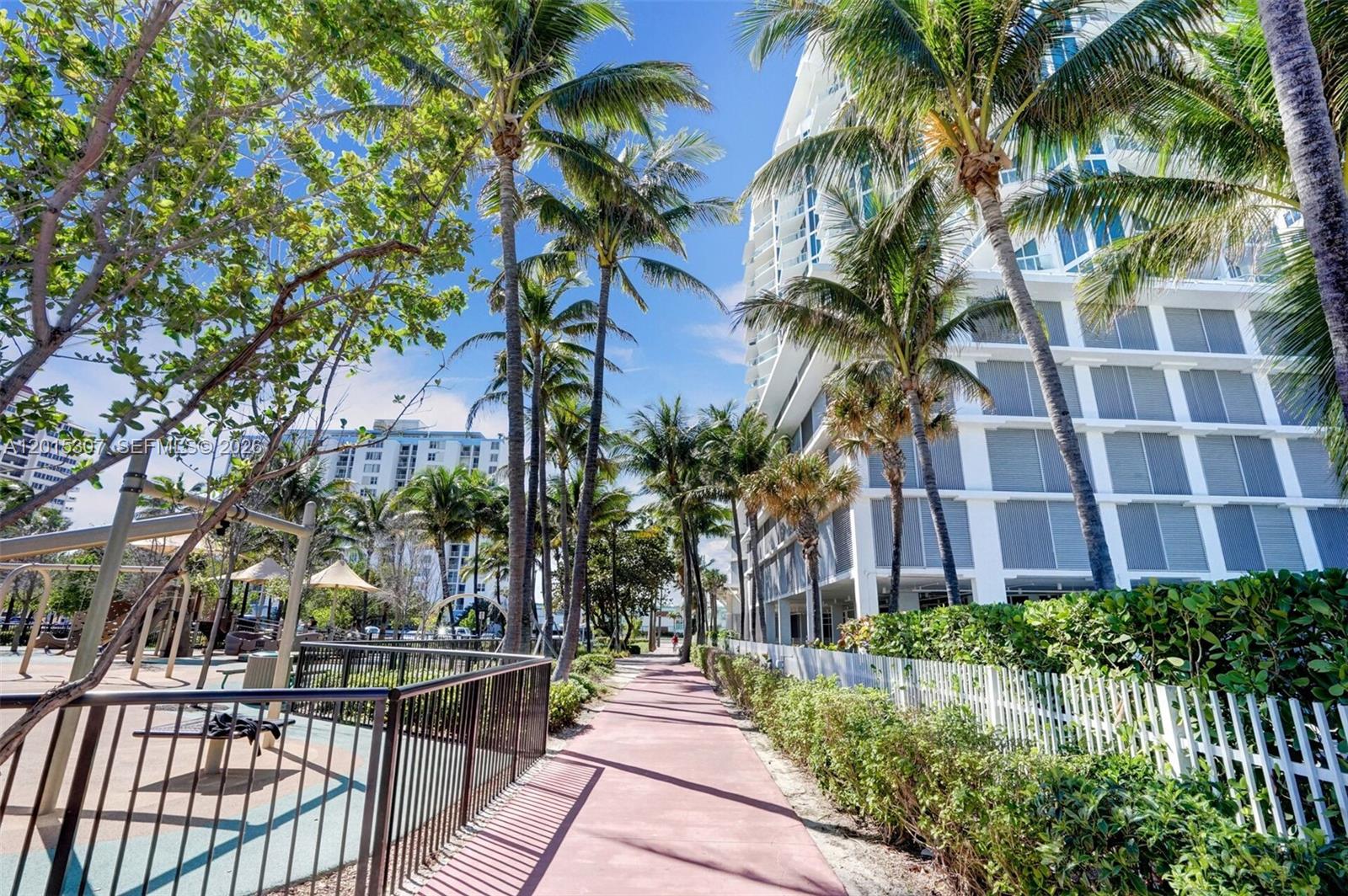 A paved walkway runs between a fenced playground and a white multi-story building with palm trees — 6515 Collins Ave # 1107, Miami Beach FL 33141, MIAMI BEACH, FL