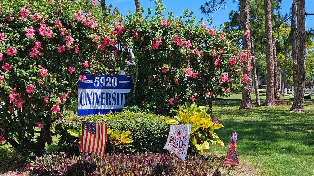 A sign reading 5920 University sits in front of a flowering bush with American flags — 5920 80TH STREET N UNIT 308, ST PETERSBURG, FL