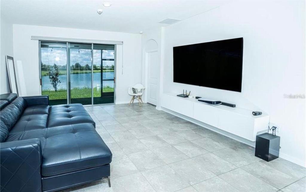 A living room with a large dark sofa, wall-mounted television, and sliding glass doors to water — 7916 STONEBROOK CIRCLE, WESLEY CHAPEL, FL