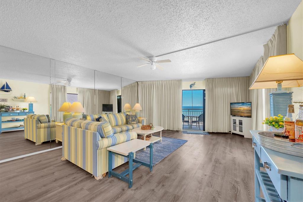 A living room features a striped sofa, mirrored wall, wood-look flooring, and a balcony view — 19440 GULF BOULEVARD UNIT 209, INDIAN SHORES, FL