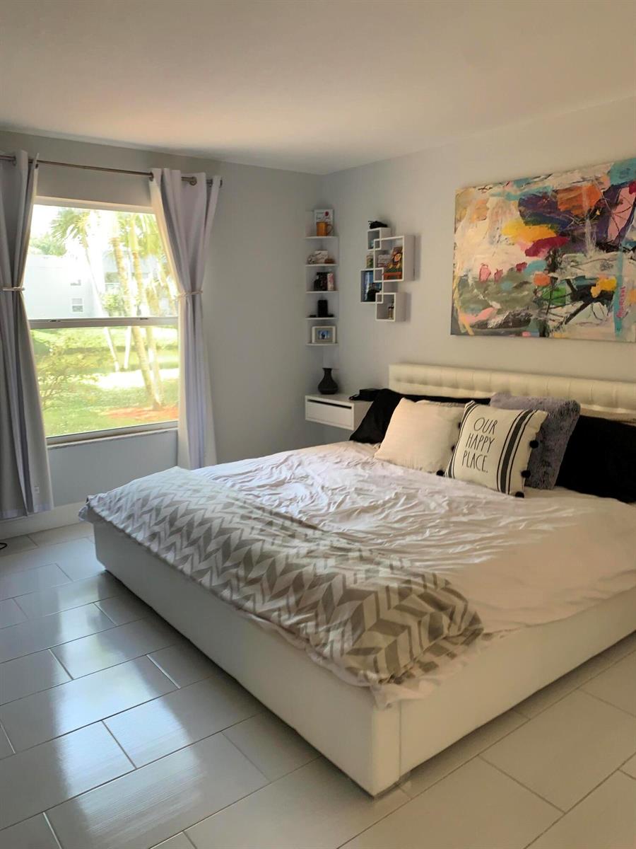A bedroom features a white bed, tiled floors, a large abstract painting, and a window — 2500 Fiore Way # 109, Delray Beach FL 33445, DELRAY BEACH, FL