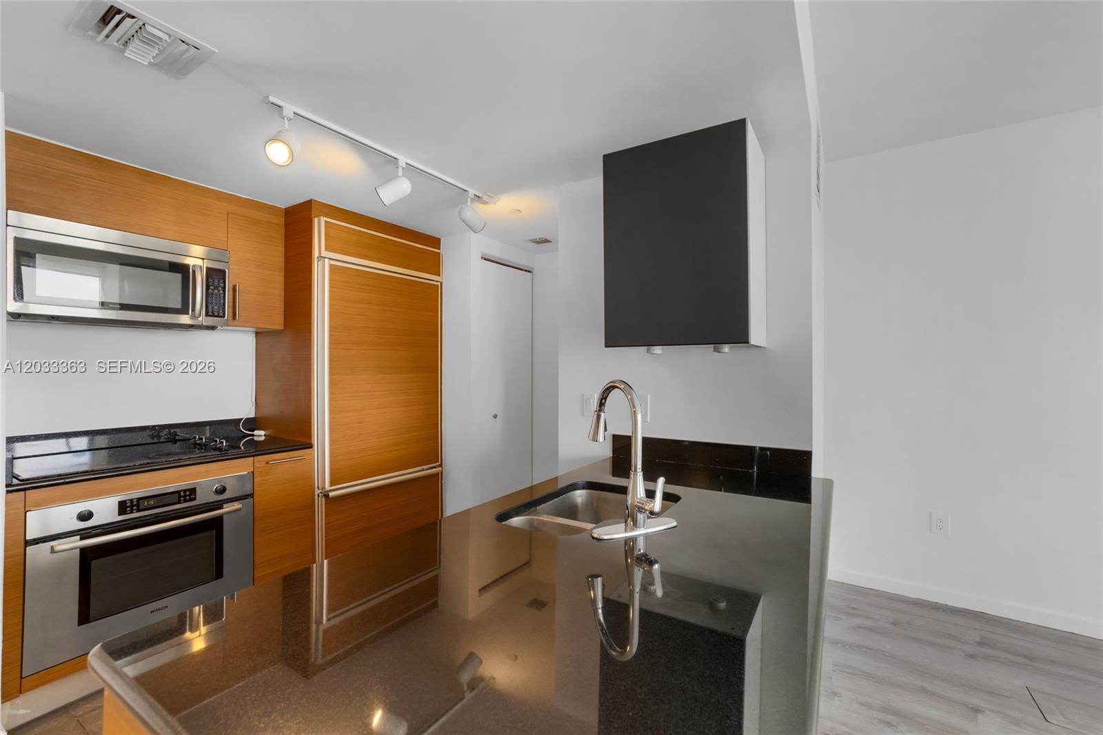 A kitchen features wood cabinetry, black countertops, stainless steel appliances, and track lighting — 50 Biscayne Blvd # 4307, Miami FL 33132, MIAMI, FL