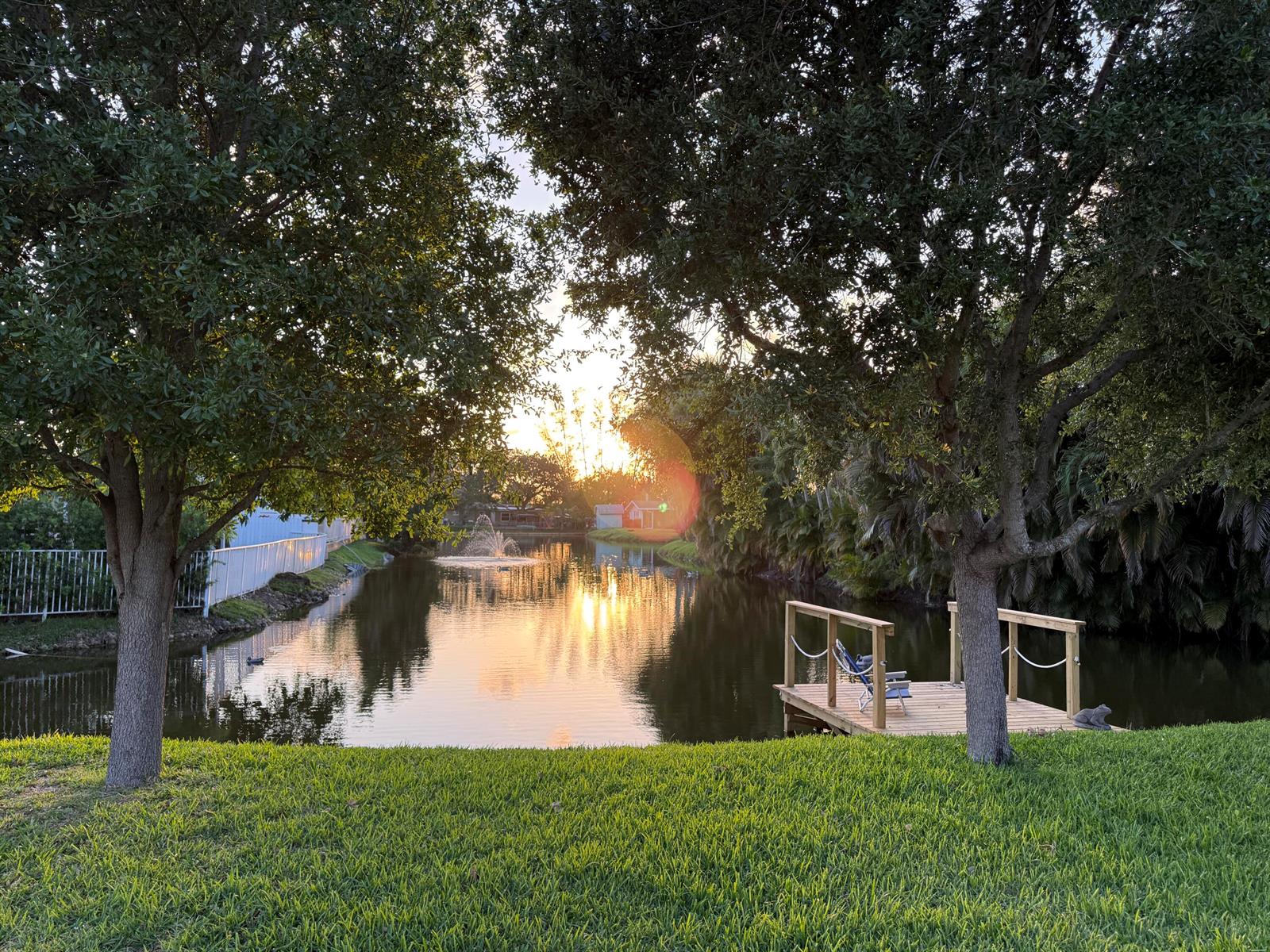 A wooden dock sits on a pond with a fountain at sunset between two trees — 1417 Willow Road, West Palm Beach FL 33406, WEST PALM BEACH, FL