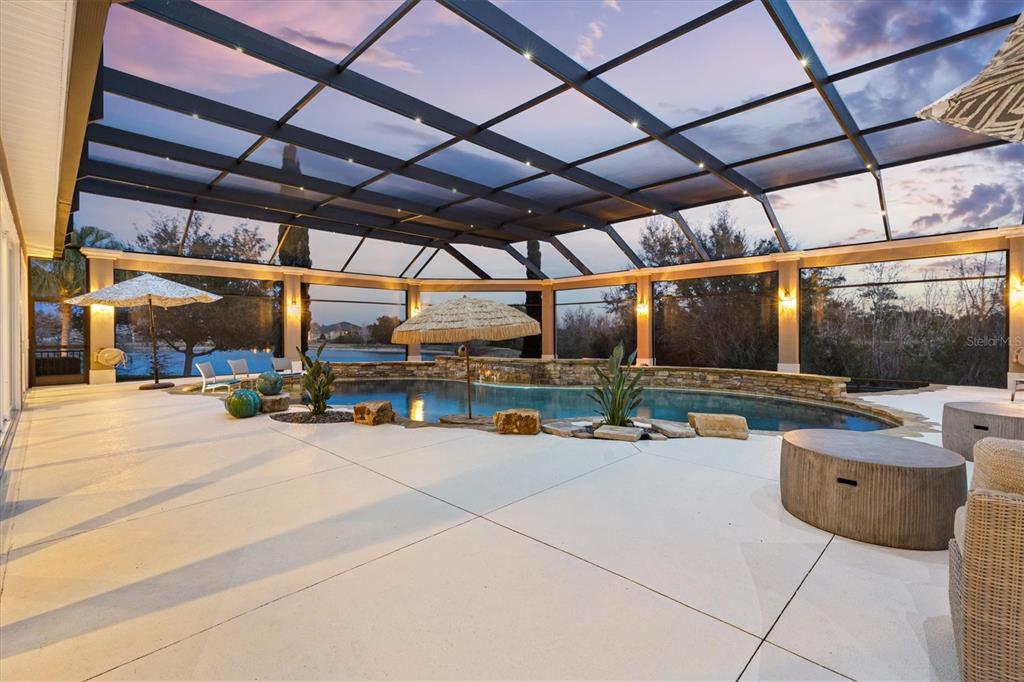 A screened-in pool patio features a stone-accented pool, lounge furniture, and a view of water — 885 IRON OAK WAY, THE VILLAGES, FL