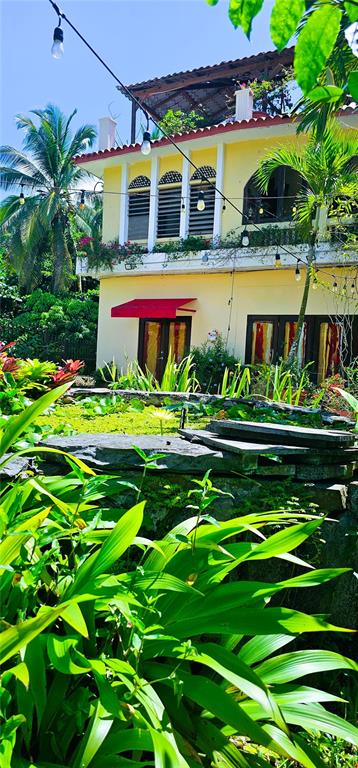 A yellow two-story building with a red awning stands behind a lush garden and pond — KM HM 0.2 ROAD 968, RIO GRANDE, PR