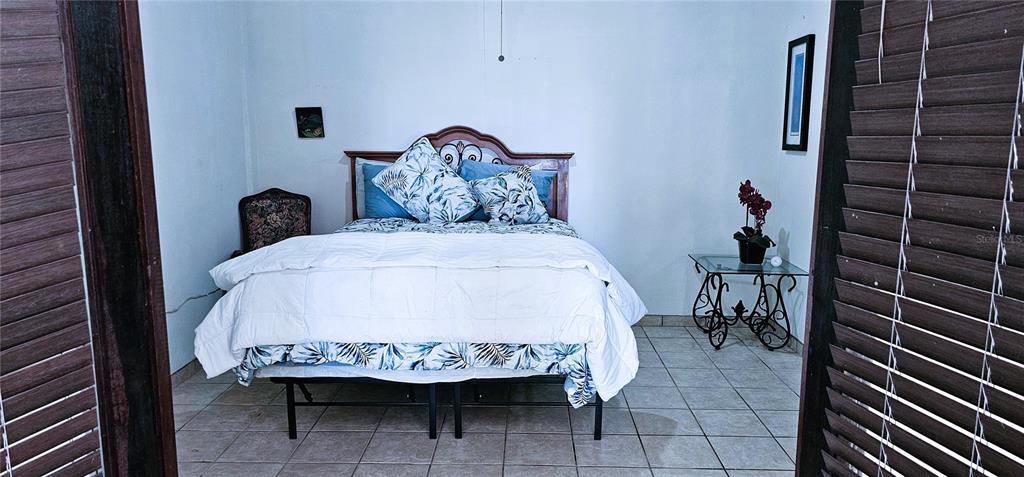 A bedroom features a bed with a patterned comforter, a small side table, and tiled flooring — KM HM 0.2 ROAD 968, RIO GRANDE, PR