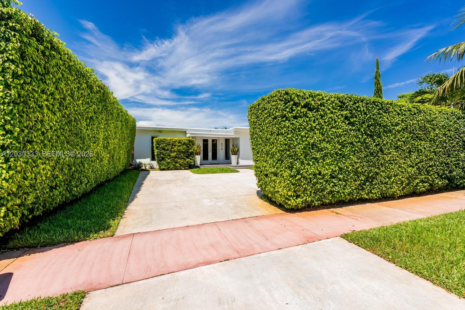 A concrete driveway leads to a white house framed by tall, manicured green hedges — 575 W 50th St, Miami Beach FL 33140, MIAMI BEACH, FL