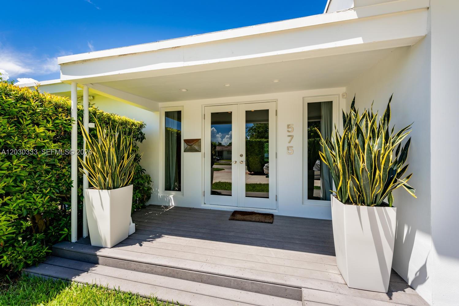 A white house entrance features double glass doors, two tall potted plants, and a wooden deck — 575 W 50th St, Miami Beach FL 33140, MIAMI BEACH, FL